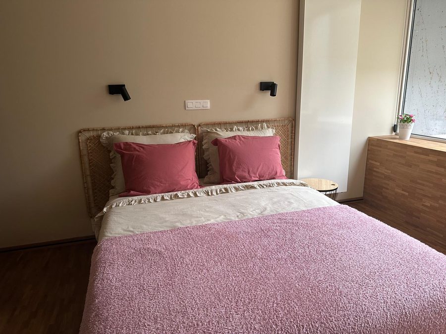 Tweepersoonsbed met roze sprei en rieten hoofdeinde bij het raam