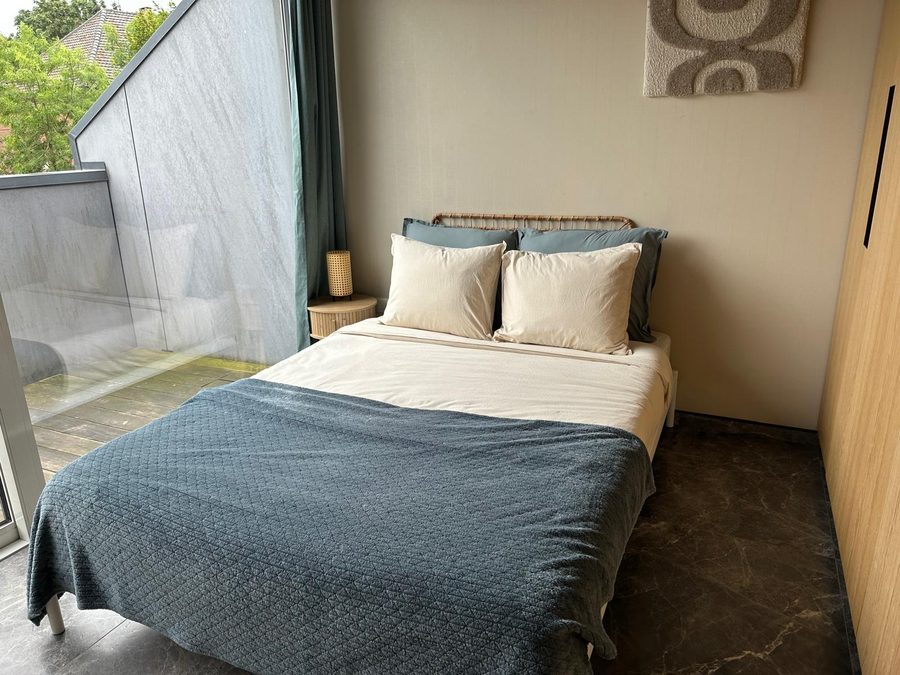 Tweepersoonsbed met blauwgroene sprei en rieten hoofdeinde naast het terrasraam